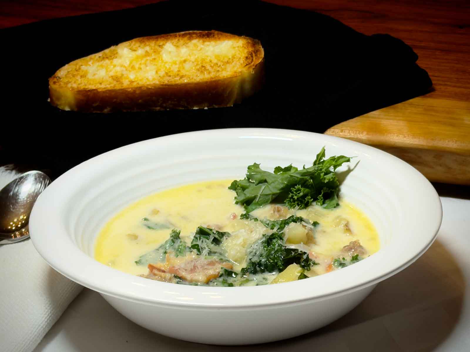 A bowl of Zuppa Toscana with sausage, potatoes, and kale in a golden broth, garnished with shaved Parmigiano Reggiano and a whole kale leaf, served with toasted bread